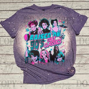 May include: Purple t-shirt with a graphic featuring characters from 90s cult classics. The design includes the text "RAISED ON 90'S CULT CLASSICS" in teal and pink. The shirt has a bleached, distressed look.