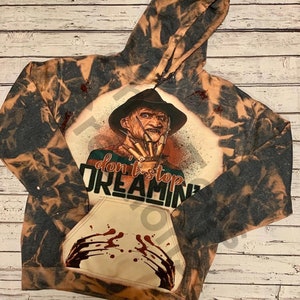 K&ouml;nnte beinhalten: Ein schwarz-beiger, mit Bleichmittel behandelter Kapuzenpullover mit einer Grafik eines vernarbten Charakters in einem Fedora mit Klingen-Handschuh. Der Text "don't stop DREAMIN'" steht &uuml;ber einer K&auml;ngurutasche mit rotem Blutspritzer-Design.