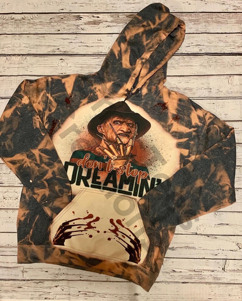 K&ouml;nnte beinhalten: Ein schwarz-beiger, mit Bleichmittel behandelter Kapuzenpullover mit einer Grafik eines vernarbten Charakters in einem Fedora mit Klingen-Handschuh. Der Text "don't stop DREAMIN'" steht &uuml;ber einer K&auml;ngurutasche mit rotem Blutspritzer-Design.