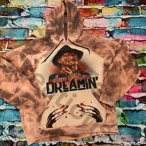 K&ouml;nnte beinhalten: Ein bleichgef&auml;rbter braun-beiger Kapuzenpullover mit einer Grafik einer Figur in einem Fedora-Hut mit Klingen an den Fingern. Der Text "don't stop DREAMIN'" ist mittig aufgedruckt, erg&auml;nzt durch blut&auml;hnliche Spritzer und Kratzspuren.