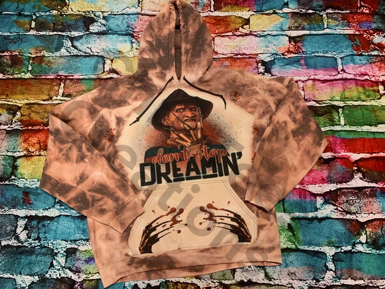K&ouml;nnte beinhalten: Ein bleichgef&auml;rbter braun-beiger Kapuzenpullover mit einer Grafik einer Figur in einem Fedora-Hut mit Klingen an den Fingern. Der Text "don't stop DREAMIN'" ist mittig aufgedruckt, erg&auml;nzt durch blut&auml;hnliche Spritzer und Kratzspuren.