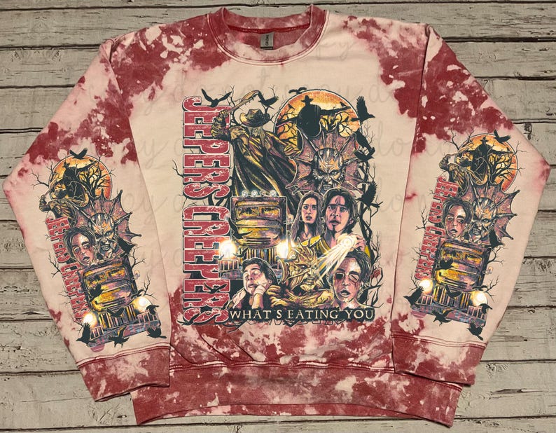 May include: A red and white tie-dye sweatshirt featuring a graphic from the movie "Jeepers Creepers." The design includes characters, a truck, and the text "What's Eating You." The sleeves also have the same graphic. The sweatshirt has a crew neck.