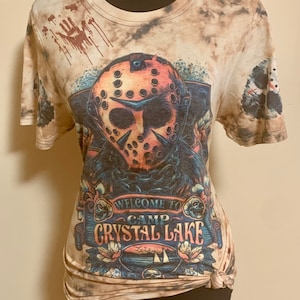 May include: A beige t-shirt featuring a graphic of a hockey mask, with the text "WELCOME TO CAMP CRYSTAL LAKE". The shirt has a tie-dye effect and a design of a handprint with blood drips.