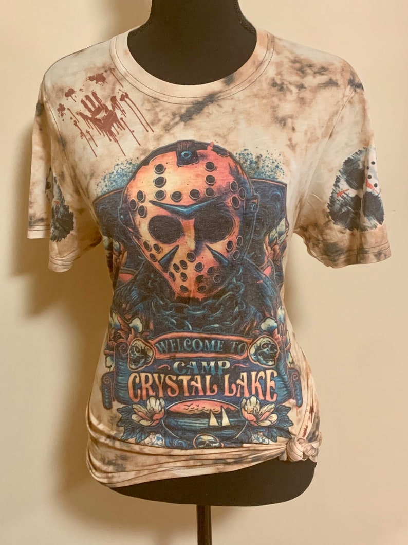 May include: A beige t-shirt featuring a graphic of a hockey mask, with the text "WELCOME TO CAMP CRYSTAL LAKE". The shirt has a tie-dye effect and a design of a handprint with blood drips.