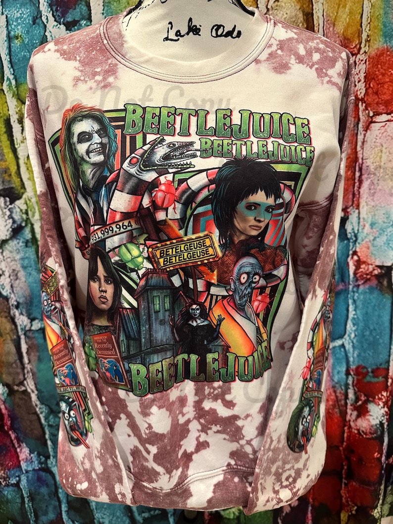 May include: A white sweatshirt with a tie-dye pattern features a colourful graphic print of characters from the film Beetlejuice. The print includes the text "Beetlejuice" and "Betelgeuse" along with images of the characters Lydia Deetz, Beetlejuice, and the Maitlands.