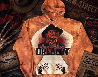 May include: A tie-dye orange hoodie featuring a graphic of a character in a fedora with bladed fingers. The text reads "don't stop DREAMIN'" above a kangaroo pocket displaying a graphic of bladed fingers. The fabric has a mottled orange tone.