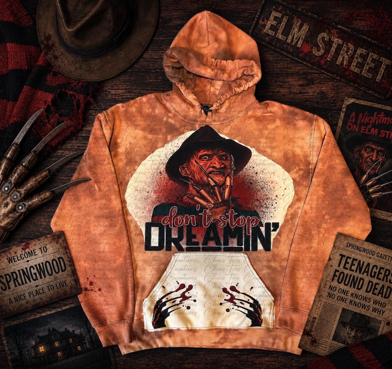 K&ouml;nnte beinhalten: Ein orangefarbener Batik-Kapuzenpullover mit einer Grafik einer Figur mit Fedora-Hut und Klingen an den Fingern. Der Text "don't stop DREAMIN'" steht &uuml;ber einer K&auml;ngurutasche mit einer Grafik von Klingen. Der Stoff hat einen fleckigen Orangeton.
