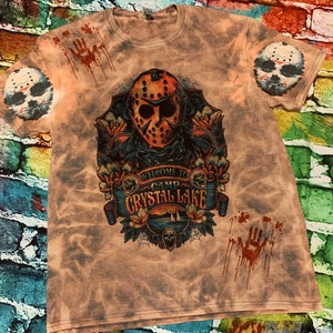 May include: A light brown t-shirt with a graphic design featuring a hockey mask, floral elements, and the text "WELCOME TO CAMP CRYSTAL LAKE." The shirt also has a splatter design and a hockey mask on each sleeve.