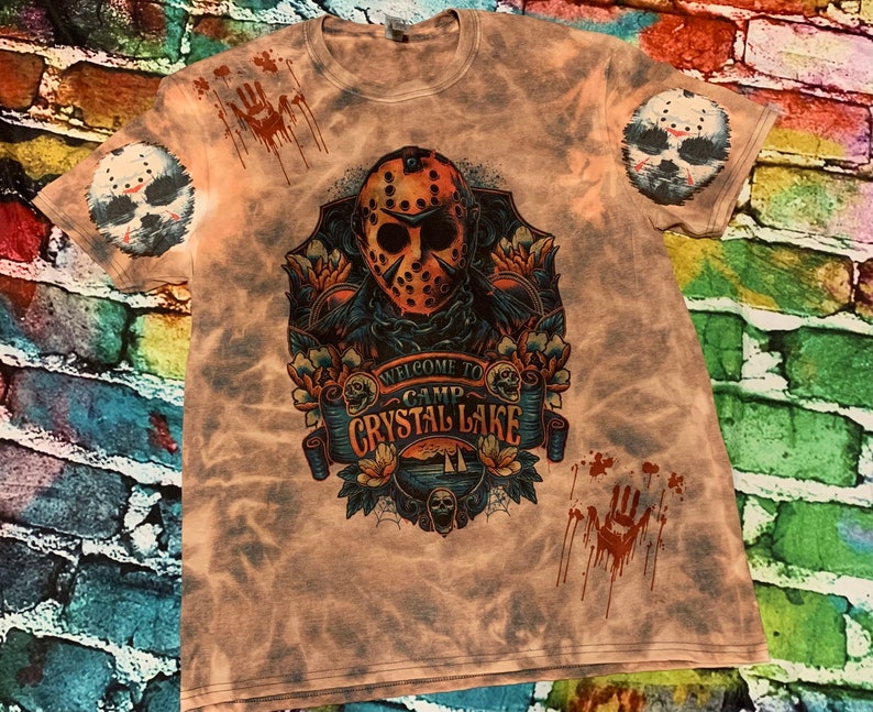 May include: A light brown t-shirt with a graphic design featuring a hockey mask, floral elements, and the text "WELCOME TO CAMP CRYSTAL LAKE." The shirt also has a splatter design and a hockey mask on each sleeve.