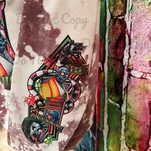 May include: White tie-dye sweatshirt featuring a graphic print of a character from the film Beetlejuice. The graphic includes a book titled "Handbook for the Recently Deceased" and the text "Betelgeuse Betelgeuse Betelgeuse".