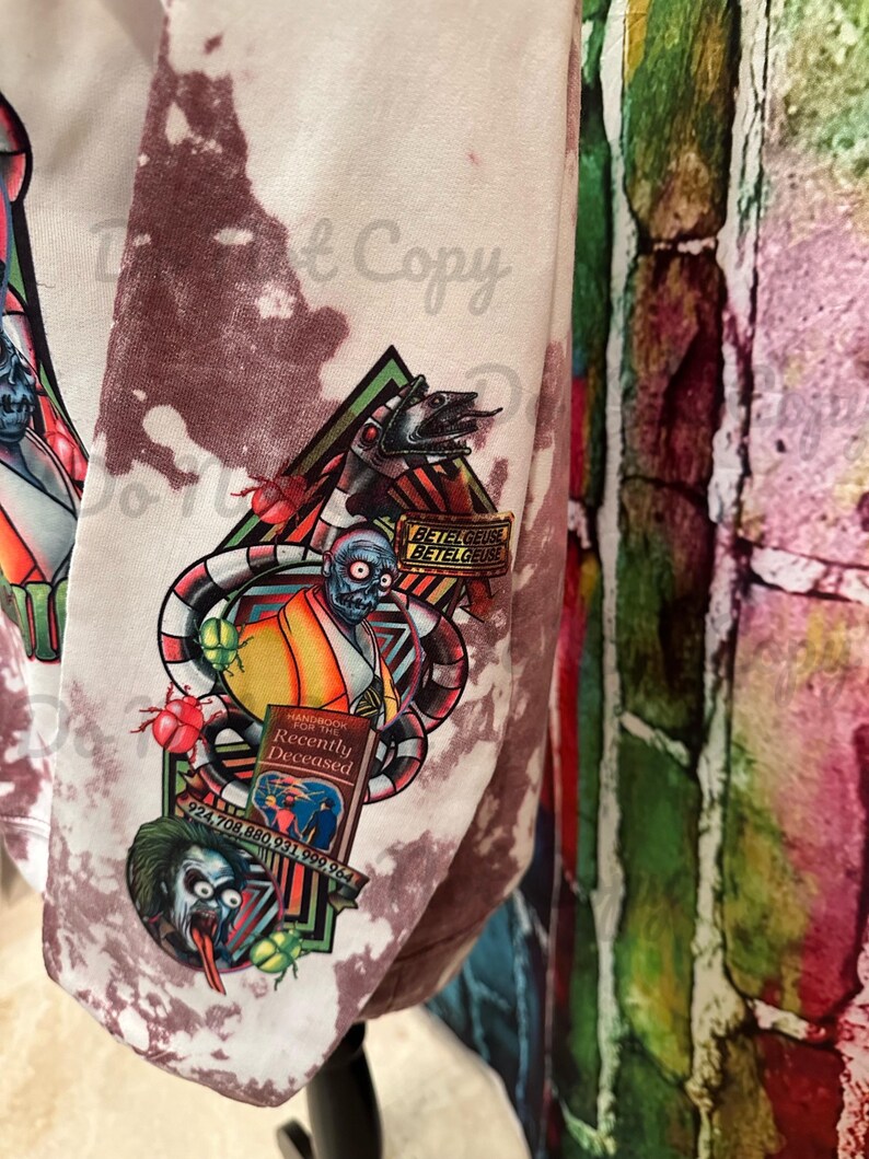 May include: White tie-dye sweatshirt featuring a graphic print of a character from the film Beetlejuice. The graphic includes a book titled "Handbook for the Recently Deceased" and the text "Betelgeuse Betelgeuse Betelgeuse".