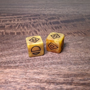 May include: Two yellow-orange dice with black geometric symbols. One die features a diamond and a half-circle with a line, while the other has a diamond and a series of angled lines. The dice are set against a wooden background.