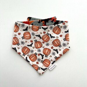 May include: A Halloween-themed dog bandana with a white background and an orange pumpkin pattern. The bandana also features black bats, spiderwebs, and candy corn. The reverse side has a pattern of gray pumpkins and orange and black accents. The label reads "kiomi paws."