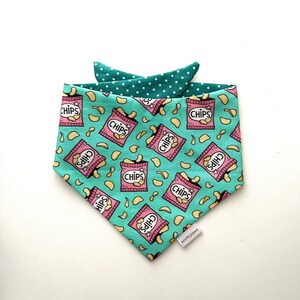 May include: A turquoise blue bandana with a pink and white chip bag pattern. The bandana has a white polka dot trim and the text "kiomi paws" is printed on the bottom right corner.