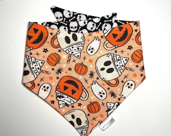 May include: Orange and black Halloween-themed dog bandana with a pattern of ghosts, pumpkins, and mugs of hot chocolate. The bandana has a black and white skull pattern on the back and a "Nomi Paws" label.