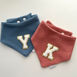 Peut inclure: Deux bavoirs pour bébé avec une texture gaufré. Un bavoir est bleu avec une lettre Y blanche avec une bordure dorée pailletée. L'autre bavoir est rose avec une lettre K blanche avec une bordure dorée pailletée. Les deux bavoirs portent le texte "korm paws".