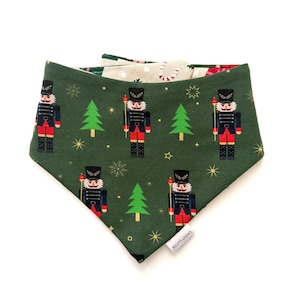 May include: A green dog bandana with a Christmas theme. The bandana features a pattern of nutcrackers, Christmas trees, and snowflakes. The reverse side has a beige and red pattern. The brand name "kiomi paws" is visible.