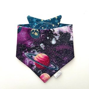 May include: A purple, black, and teal dog bandana with a space theme. The bandana features planets, stars, and a galaxy print. The bandana has a white tag that says "kosmi paws".