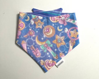 May include: A blue dog bandana with a repeating pattern of stars, moons, wands, and hearts in pastel colors. The bandana has a contrasting blue and purple striped collar. The tag reads "kiomi paws".