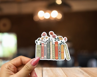 May include: A sticker featuring a stack of vintage books with flowers growing from the top. The books have different coloured spines and are arranged in a row. The flowers are pink, orange, and yellow.