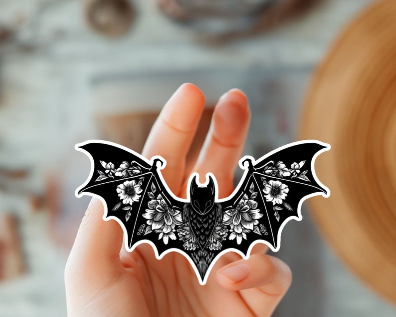 May include: Black and white sticker of a bat with floral details. The bat is flying with its wings spread out and has a floral pattern on its wings. The sticker is on a hand.