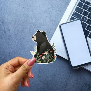 Puede incluir: Una pegatina de un oso negro con un dise&ntilde;o floral. El oso est&aacute; sentado en un campo de flores con hojas verdes. La pegatina est&aacute; sobre un fondo gris.