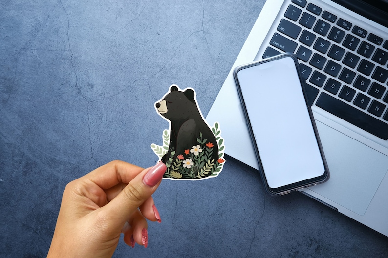Puede incluir: Una pegatina de un oso negro con un dise&ntilde;o floral. El oso est&aacute; sentado en un campo de flores con hojas verdes. La pegatina est&aacute; sobre un fondo gris.