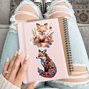 May include: A pink notebook with a spiral binding and two fox stickers. One sticker features a fox surrounded by flowers, and the other shows a fox with closed eyes surrounded by flowers. The notebook is held on a person's lap, with ripped jeans and black sneakers visible.