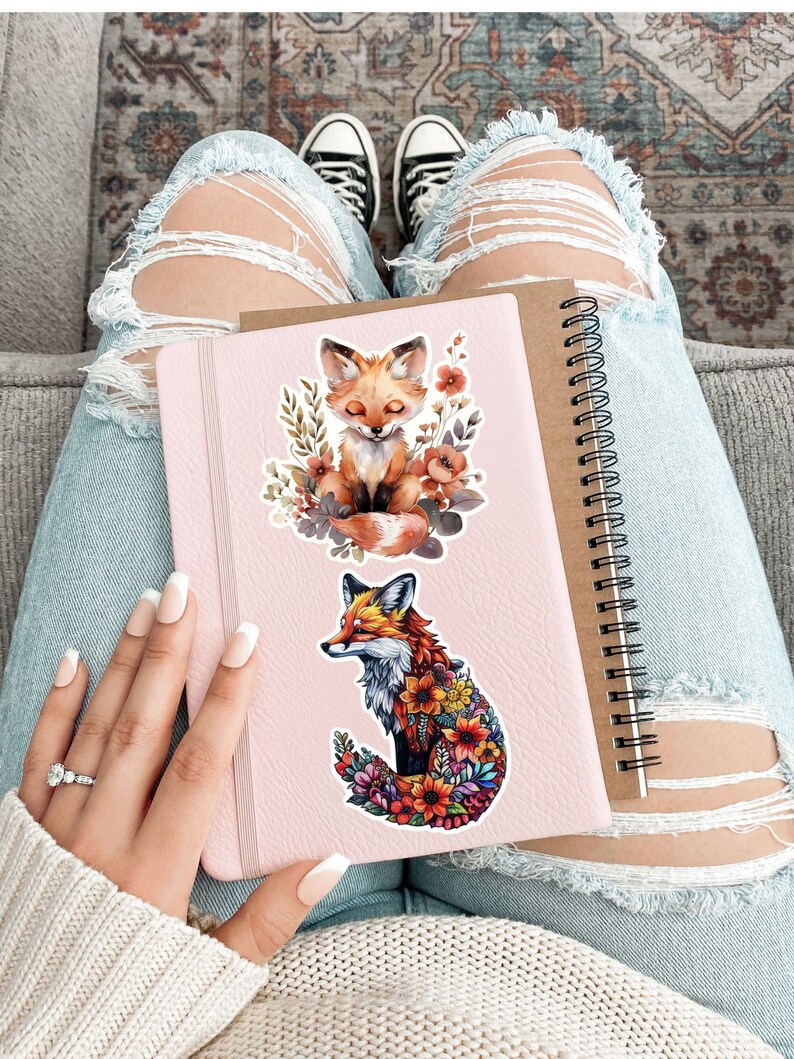 May include: A pink notebook with a spiral binding and two fox stickers. One sticker features a fox surrounded by flowers, and the other shows a fox with closed eyes surrounded by flowers. The notebook is held on a person's lap, with ripped jeans and black sneakers visible.