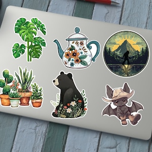 Puede incluir: Un conjunto de seis pegatinas con varios dise&ntilde;os, que incluyen una tetera con patrones florales, un oso negro sentado en un campo de flores, un murci&eacute;lago con un sombrero de vaquero, un grupo de plantas en macetas y una silueta de Bigfoot en una escena de bosque.