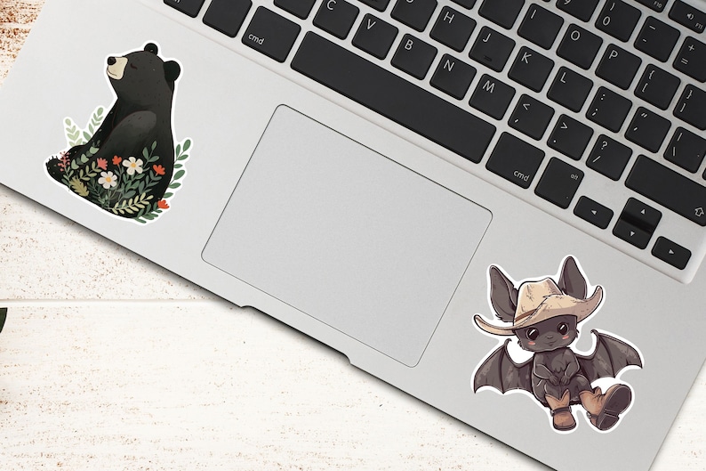 Puede incluir: Dos pegatinas en un port&aacute;til. Una pegatina presenta un oso negro sentado en un lecho de flores. La otra pegatina presenta un murci&eacute;lago de dibujos animados con un sombrero de vaquero y botas.