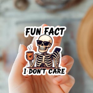 May include: A sticker featuring a cartoon skeleton wearing sunglasses, holding a coffee mug with a heart and a smartphone. The text on the sticker reads "FUN FACT I DON'T CARE". The sticker is held in a hand.