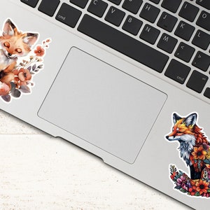 May include: Two stickers featuring fox illustrations. One shows a sleeping fox surrounded by flowers in shades of orange, brown, and purple. The other depicts a fox with a floral design in vibrant colors. Both are placed on a laptop.