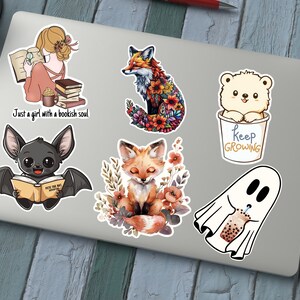 May include: A collection of colorful stickers featuring whimsical illustrations. Designs include a girl reading with the text "Just a girl with a bookish soul", a fox with floral details, a bear in a cup with "Keep Growing", a bat reading, a fox with flowers, and a ghost with boba.