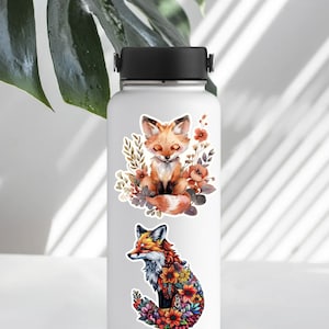 May include: White water bottle with two fox illustrations. One fox is sitting with closed eyes, surrounded by flowers. The other fox is made of colorful flowers. The bottle has a black lid and handle.