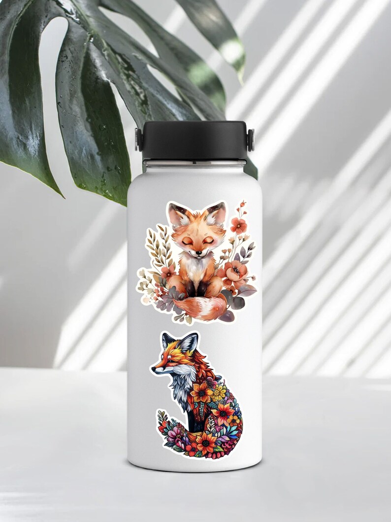 May include: White water bottle with two fox illustrations. One fox is sitting with closed eyes, surrounded by flowers. The other fox is made of colorful flowers. The bottle has a black lid and handle.