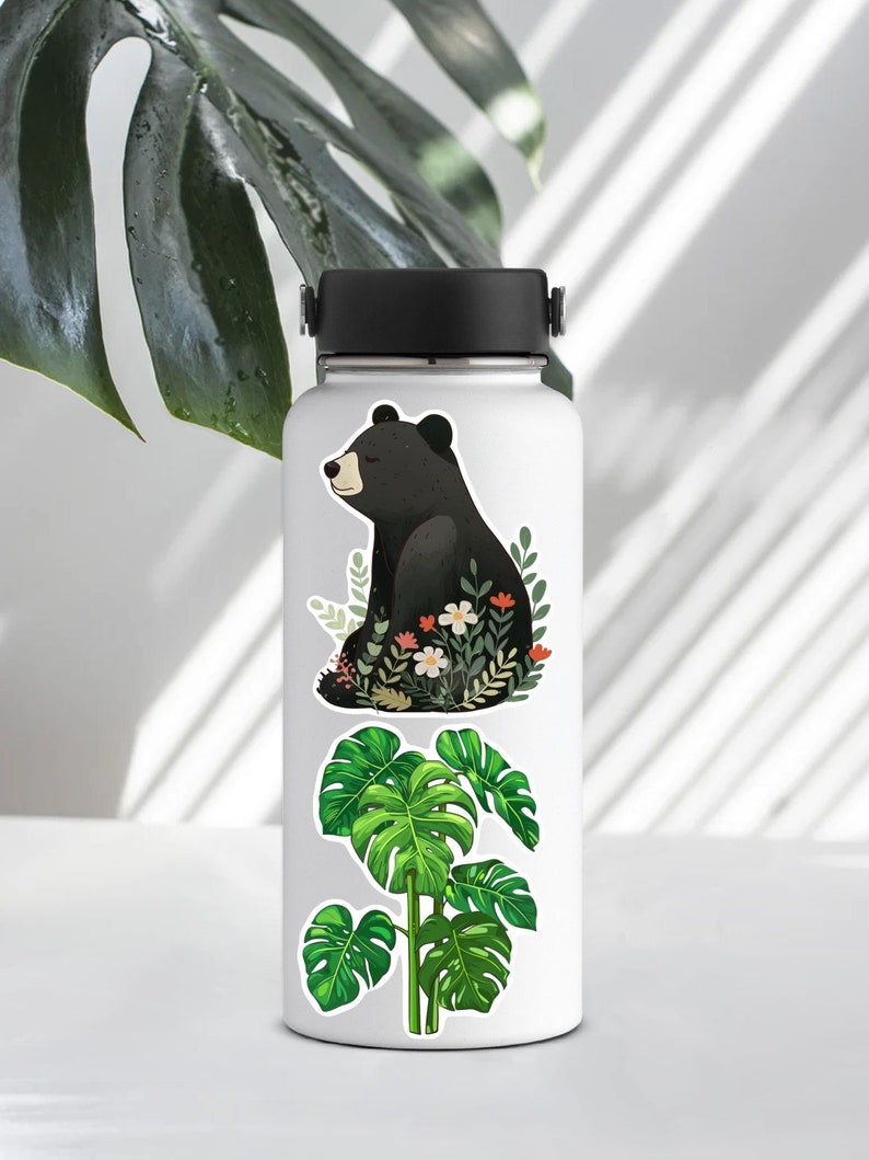 Puede incluir: Botella de agua blanca con tres pegatinas. La pegatina superior presenta un oso negro sentado en un dise&ntilde;o floral verde y blanco. La pegatina del medio presenta una planta de monstera verde. La pegatina inferior presenta una planta de monstera verde.
