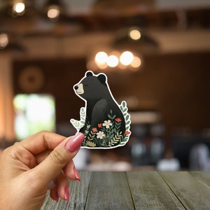 Puede incluir: Una pegatina de un oso negro con un dise&ntilde;o floral. El oso est&aacute; sentado en un lecho de hojas verdes y flores blancas y rojas.