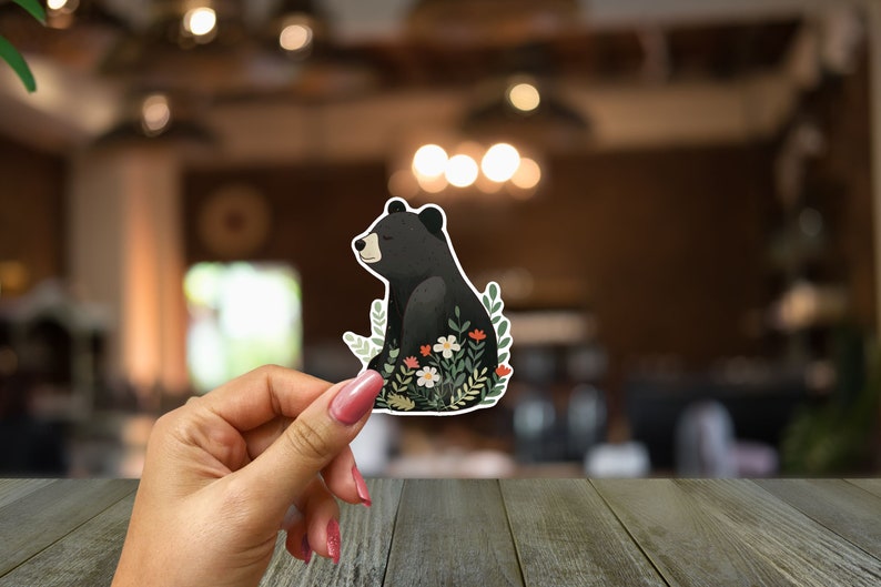Puede incluir: Una pegatina de un oso negro con un dise&ntilde;o floral. El oso est&aacute; sentado en un lecho de hojas verdes y flores blancas y rojas.