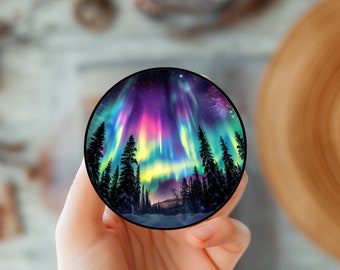 May include: A round sticker with a colorful aurora borealis scene. The aurora is green, purple, pink, and yellow. The scene is set against a dark sky with stars. There are dark silhouette trees in the foreground.