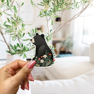 Puede incluir: Una pegatina de un oso negro con un dise&ntilde;o floral. El oso est&aacute; sentado en un campo de flores con hojas verdes. La pegatina est&aacute; siendo sostenida por una mano.