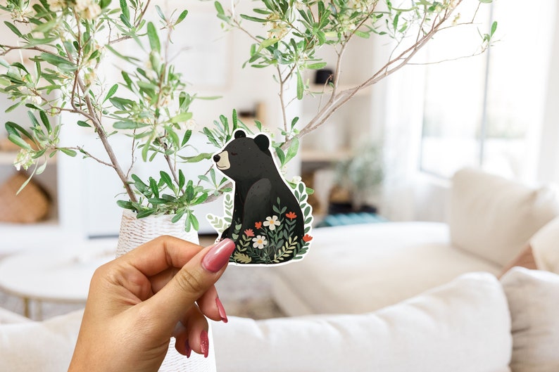 Puede incluir: Una pegatina de un oso negro con un dise&ntilde;o floral. El oso est&aacute; sentado en un campo de flores con hojas verdes. La pegatina est&aacute; siendo sostenida por una mano.