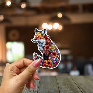 May include: A colorful sticker featuring a fox design. The fox is depicted with a vibrant mix of orange, red, and white fur, and its body is filled with various flowers. The sticker is held against a blurred background.