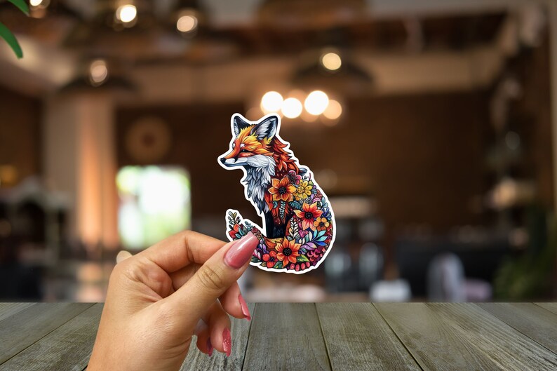 May include: A colorful sticker featuring a fox design. The fox is depicted with a vibrant mix of orange, red, and white fur, and its body is filled with various flowers. The sticker is held against a blurred background.