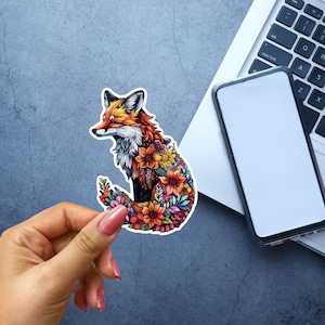 May include: A colorful sticker featuring a fox design. The fox is depicted with orange, red, and white fur, and its body is filled with vibrant flowers. The sticker is held by a hand, with a laptop and phone in the background.