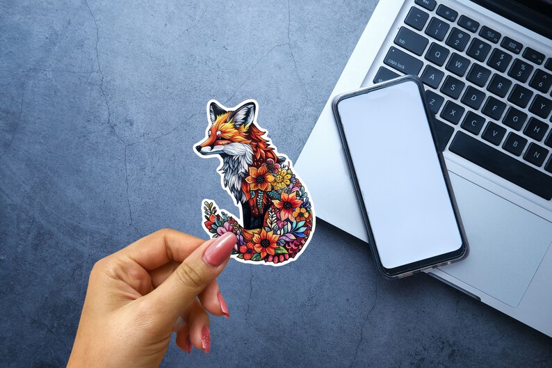 May include: A colorful sticker featuring a fox design. The fox is depicted with orange, red, and white fur, and its body is filled with vibrant flowers. The sticker is held by a hand, with a laptop and phone in the background.