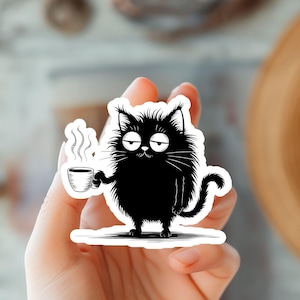 May include: A black and white sticker featuring a cartoon cat with a grumpy expression, holding a steaming coffee cup. The cat has fluffy fur and a curved tail. The sticker is on a white background.