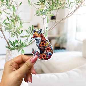 May include: A colorful sticker featuring a fox with a floral design. The fox has orange, brown, and white fur, and its body is filled with various flowers in shades of red, orange, yellow, and purple. The sticker is held in a hand.