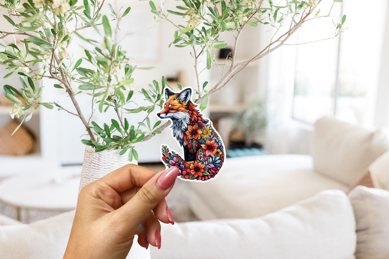May include: A colorful sticker featuring a fox with a floral design. The fox has orange, brown, and white fur, and its body is filled with various flowers in shades of red, orange, yellow, and purple. The sticker is held in a hand.