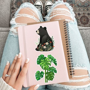 Puede incluir: Una libreta rosa con una pegatina de un oso negro y una planta de monstera verde. La libreta tiene una encuadernaci&oacute;n en espiral.